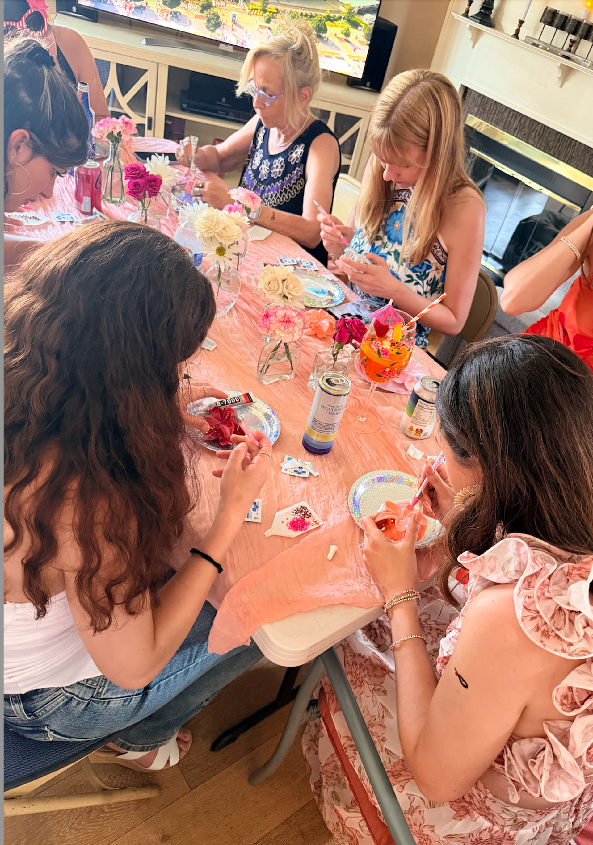 Women and girls at a DIY craft party around a pink-covered table in a living room, decorating small projects with beads, paint, drinks and fresh flowers