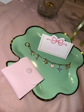 Mint-green scalloped plate with gold rim holding a charm bracelet, small pink velvet pouch, and a pink bow place card on a pale pink tablecloth — cute party favor table setting.