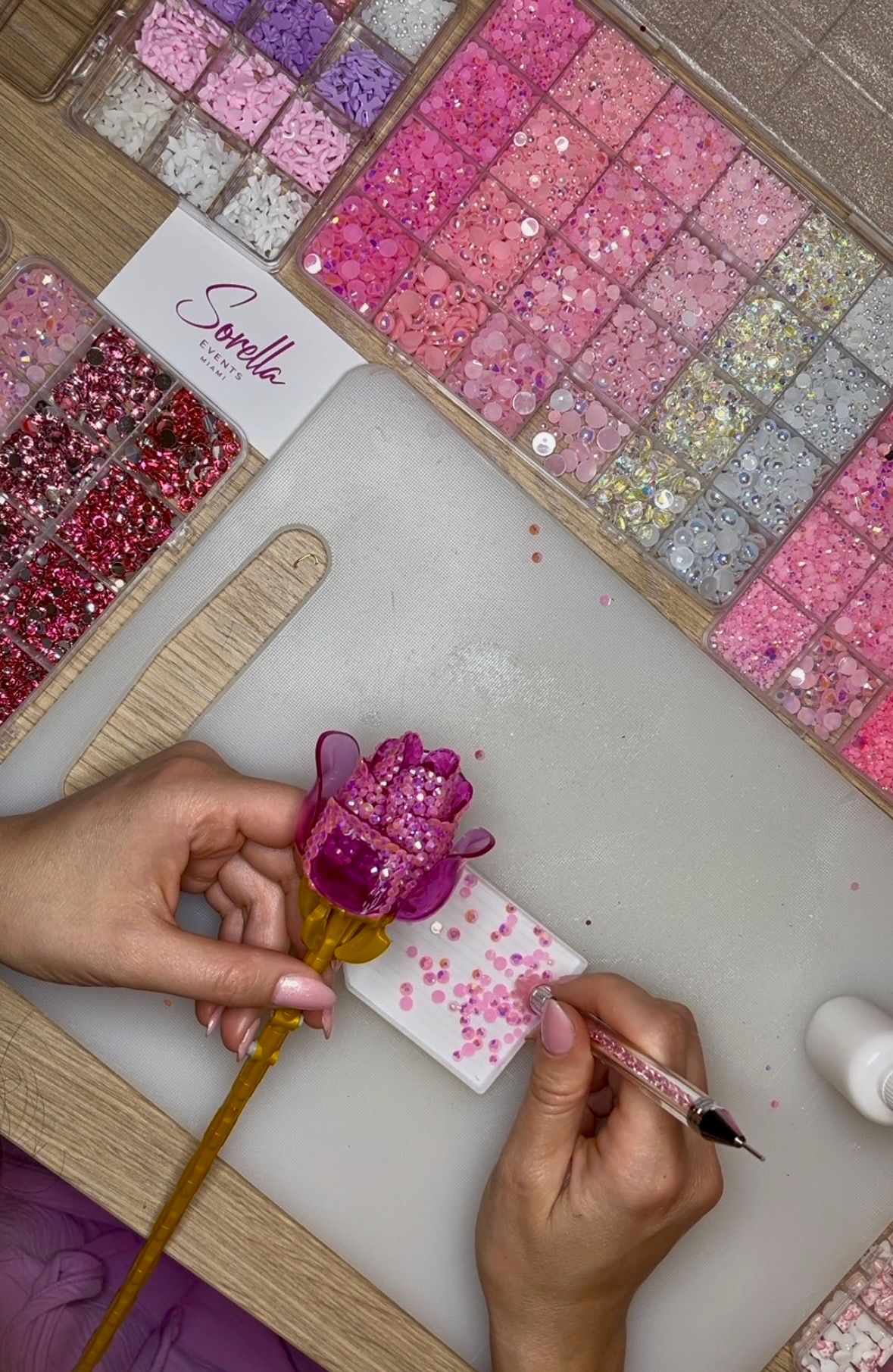Hands decorating a translucent pink resin rose on a yellow stem with pink rhinestones using a crystal-tipped picker, surrounded by organized trays of pink, purple and iridescent sequins and beads on a craft mat