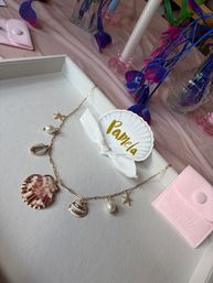 Beachy gold charm necklace with shell, pearl and starfish on a white jewelry tray, gold-lettered shell name tag tied with a white ribbon, pink suede pouch and vivid blue-purple faux flowers on a pink tablecloth.