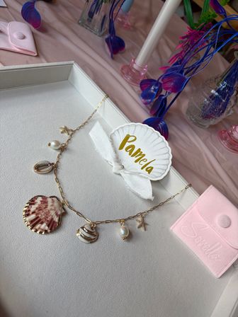 Beachy gold charm necklace with shell, pearl and starfish on a white jewelry tray, gold-lettered shell name tag tied with a white ribbon, pink suede pouch and vivid blue-purple faux flowers on a pink tablecloth.