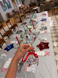 Hand holding a decorated martini glass covered in red, blue and silver star stickers with a red ribbon, hovering over a long indoor craft table set with star-shaped plates, red and blue candles and chairs for a patriotic-themed party.