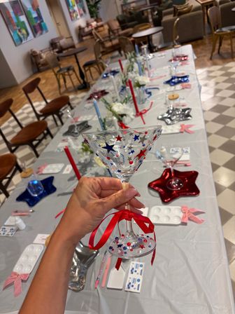 Hand holding a decorated martini glass covered in red, blue and silver star stickers with a red ribbon, hovering over a long indoor craft table set with star-shaped plates, red and blue candles and chairs for a patriotic-themed party.