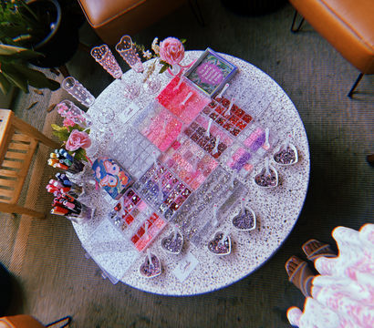 Overhead view of a round terrazzo craft table set up for DIY jewelry with clear organizers of pink, red and purple beads and rhinestones, heart-shaped scoops, glitter, markers, pink roses and champagne flutes in a cozy indoor seating area