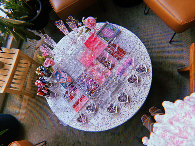 Overhead view of a round terrazzo craft table set up for DIY jewelry with clear organizers of pink, red and purple beads and rhinestones, heart-shaped scoops, glitter, markers, pink roses and champagne flutes in a cozy indoor seating area
