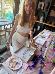 Smiling woman in a white two-piece leans over a bright jewelry-making display with gold necklaces, shell charms, pliers and colorful floral accents by a window with turquoise outdoor chairs visible