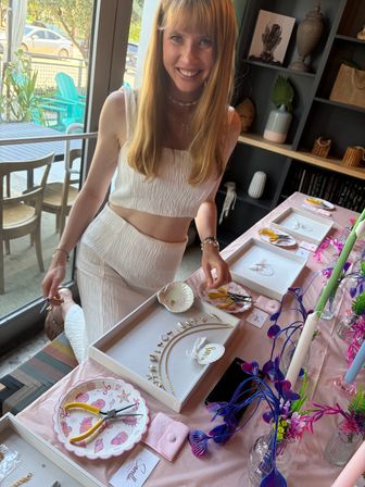 Smiling woman in a white two-piece leans over a bright jewelry-making display with gold necklaces, shell charms, pliers and colorful floral accents by a window with turquoise outdoor chairs visible