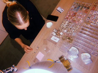 Overhead view of a DIY jewelry-making workspace: person arranging beads, charms and shells in clear divided trays on a wooden table with pliers, phone and a small heart dish.