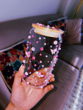 Hand holding a clear glass jar decorated with pink pearls, rhinestones and tiny butterfly stickers with a wooden lid, cozy living room couch and colorful floral pillow blurred in the background.
