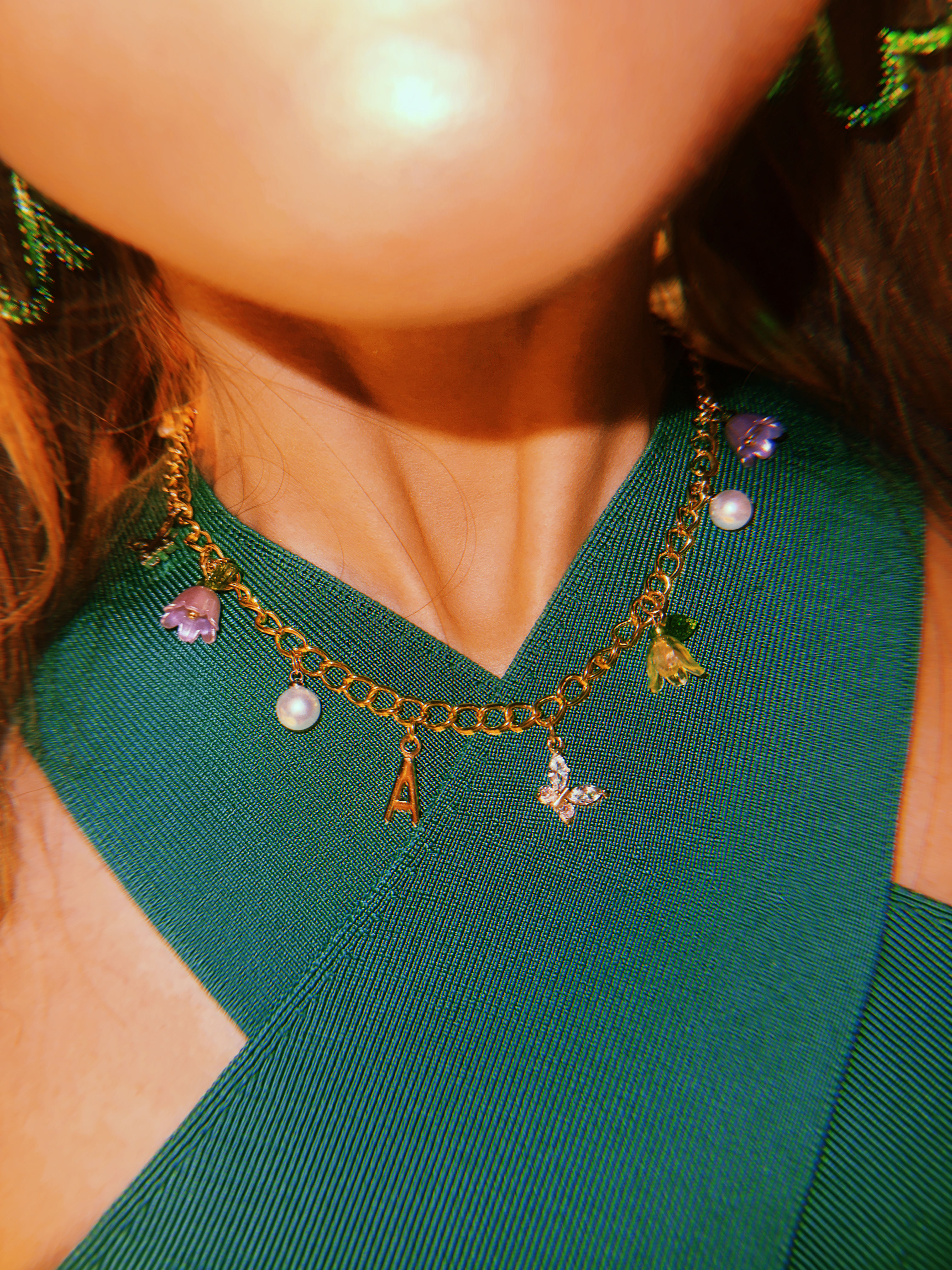 Close-up of a neckline in a green ribbed V-top wearing a gold charm chain necklace with an 'A' initial, pearl, butterfly and flower charms — fashion accessory detail