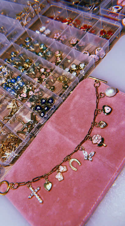 Sparkly gold charm bracelet with cross, heart, horseshoe, rose and butterfly charms on a pink velvet pouch next to a clear organizer of beads, evil-eye charms and jewelry-making findings.