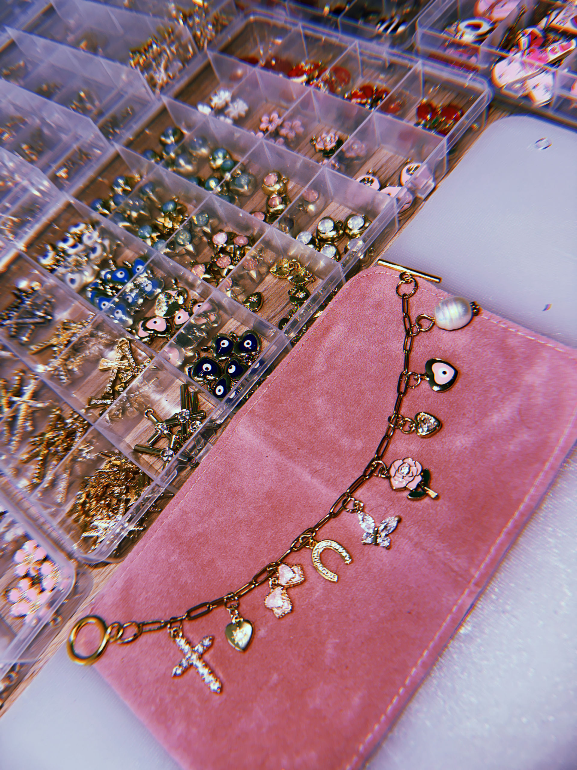 Sparkly gold charm bracelet with cross, heart, horseshoe, rose and butterfly charms on a pink velvet pouch next to a clear organizer of beads, evil-eye charms and jewelry-making findings.