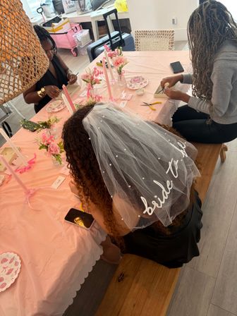 Indoor bachelorette/bridal shower scene with bride-to-be veil at a pink-decorated table as friends craft floral favors, candles, and place settings in a bright home setting