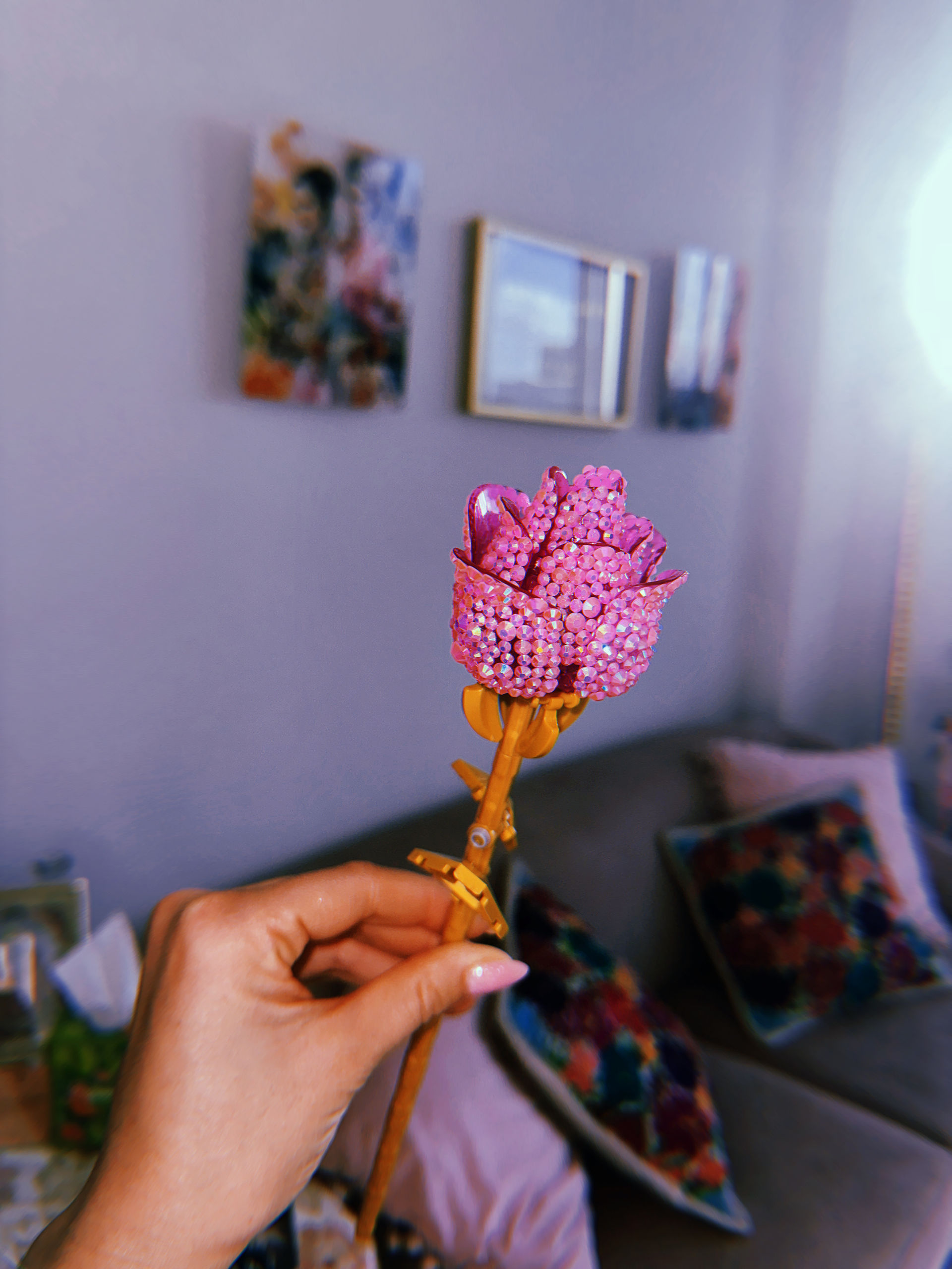 Hand holding a sparkly pink beaded rose on a gold stem against a cozy living room backdrop with colorful throw pillows and framed wall art
