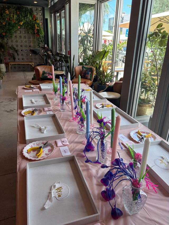 Sunlit plant-filled cafe interior with a long pink-covered table set for a pastel candle craft workshop, glass vases, tools and gift boxes by large windows