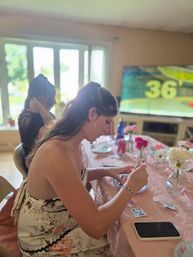Young woman in a floral sundress crafting at a pink-covered dining table in a sunlit living room, vases of pink and white flowers, smartphone and art supplies on the table, blurred TV and window in the background.