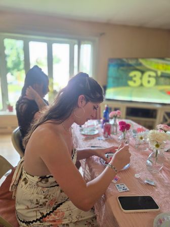 Young woman in a floral sundress crafting at a pink-covered dining table in a sunlit living room, vases of pink and white flowers, smartphone and art supplies on the table, blurred TV and window in the background.