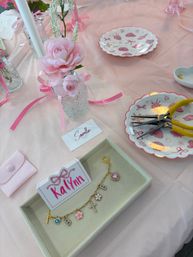 Pink party DIY bracelet station with faux pink roses in glass vases, a charm bracelet displayed in a tray, yellow-handled pliers on scalloped seashell plates, and a soft pink tablecloth.