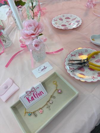Pink party DIY bracelet station with faux pink roses in glass vases, a charm bracelet displayed in a tray, yellow-handled pliers on scalloped seashell plates, and a soft pink tablecloth.