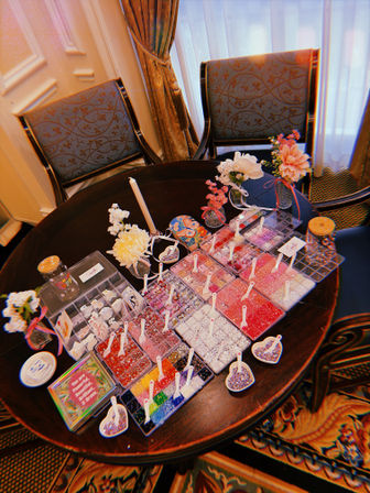 Overhead view of a round wooden table set up as a colorful DIY bead station with organized trays of beads and rhinestones, tiny scoops, heart bowls, small floral vases and a candle, flanked by two patterned upholstered chairs and draped curtains.