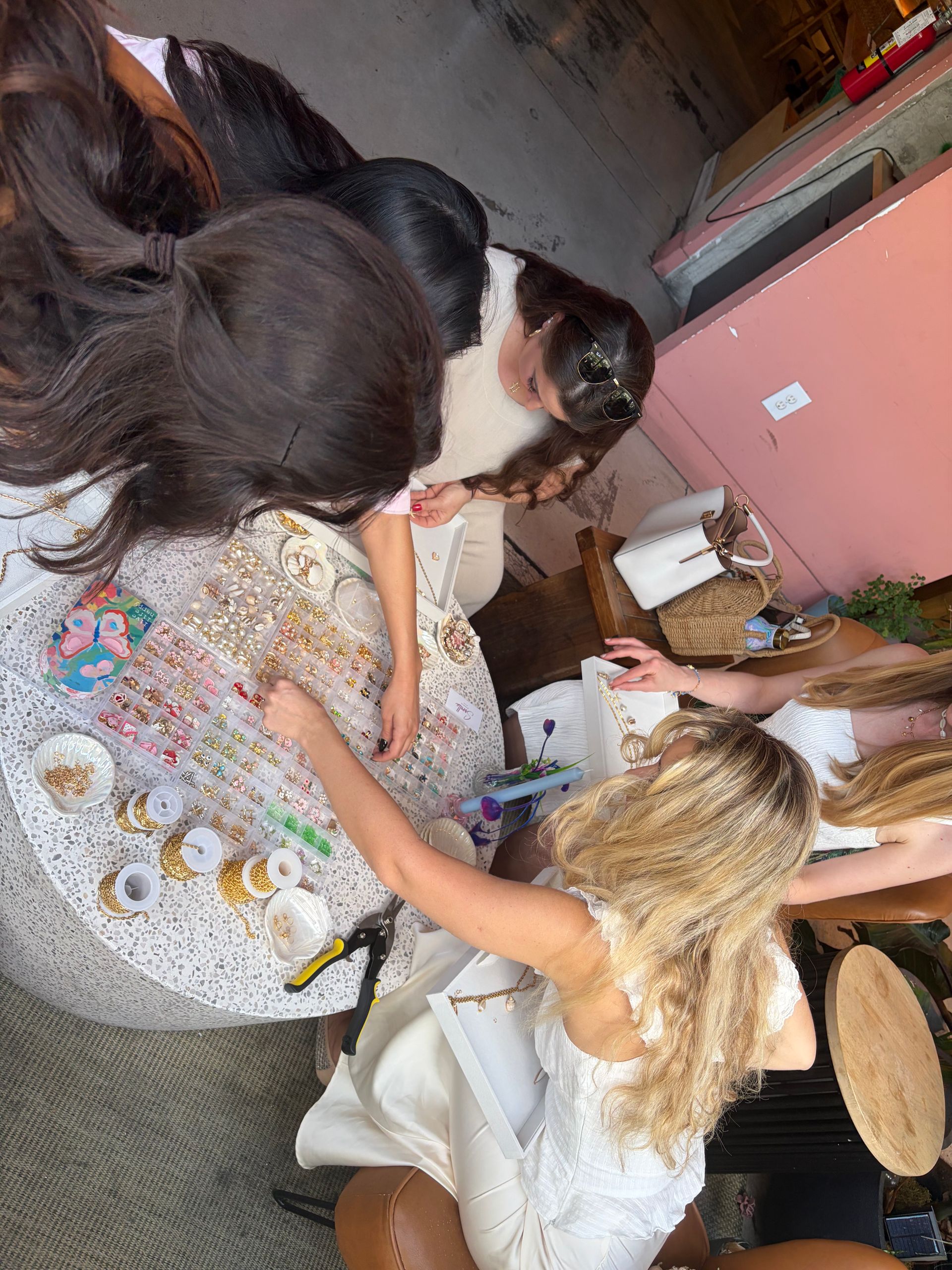 Group of women at a jewelry beading party assembling handmade bracelets and necklaces — trays of colorful charms, gold chain spools, pliers and handbags on a pink-walled table.