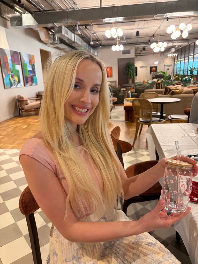 Smiling blonde woman holding a floral-decorated glass jar with a straw in a modern café lounge with globe pendant lights, cozy seating and colorful wall art.