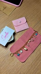 Top-down flatlay on a wood tabletop showing a pink suede jewelry roll opened to reveal a gold charm bracelet with starfish, pearl, flower and heart charms, a matching pink snap pouch, a pale business card and a pink smartphone corner