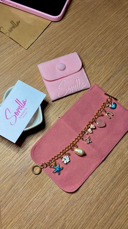 Top-down flatlay on a wood tabletop showing a pink suede jewelry roll opened to reveal a gold charm bracelet with starfish, pearl, flower and heart charms, a matching pink snap pouch, a pale business card and a pink smartphone corner
