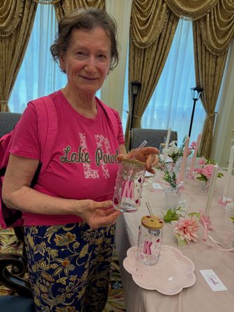Smiling person in a pink shirt holding a decorated clear jar with a straw at a pink-themed craft table with candles, flowers and pale-gold drapery in an elegant indoor event room