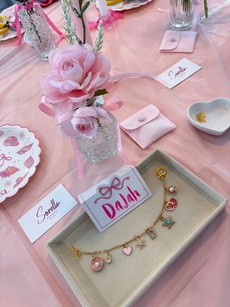 Pretty pink party table with a faux peony in a cut-glass vase tied with ribbon, scalloped seashell plates, small pink favor pouches, a heart dish with gold beads, and a tray displaying a gold charm bracelet and pink place card.