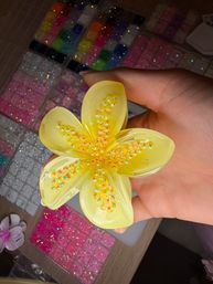 Hand holding a bright yellow resin flower studded with iridescent sequins above organized trays of colorful glitter beads and craft supplies on a table