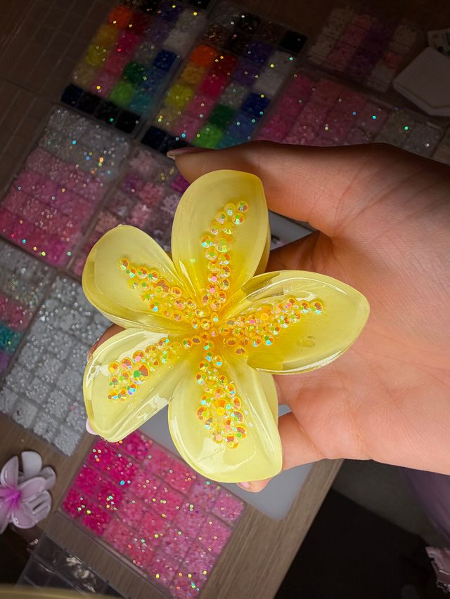Hand holding a bright yellow resin flower studded with iridescent sequins above organized trays of colorful glitter beads and craft supplies on a table