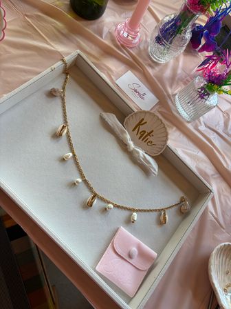Stylish flat-lay of a jewelry display on a pale pink table: white tray holding a gold chain necklace with cowrie shells and pearls, a shell-shaped name tag tied with ribbon, a soft pink suede pouch, and nearby glass vases of pink and purple flowers plus a pink candle.