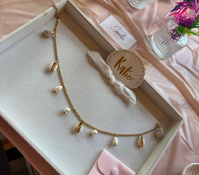 Stylish flat-lay of a jewelry display on a pale pink table: white tray holding a gold chain necklace with cowrie shells and pearls, a shell-shaped name tag tied with ribbon, a soft pink suede pouch, and nearby glass vases of pink and purple flowers plus a pink candle.