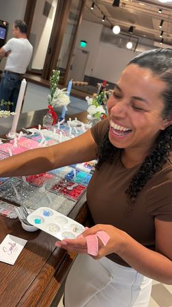 Laughing woman selecting colorful beads and sequins from organized trays and a paint palette at a bright DIY craft/bead bar with flowers on the counter.