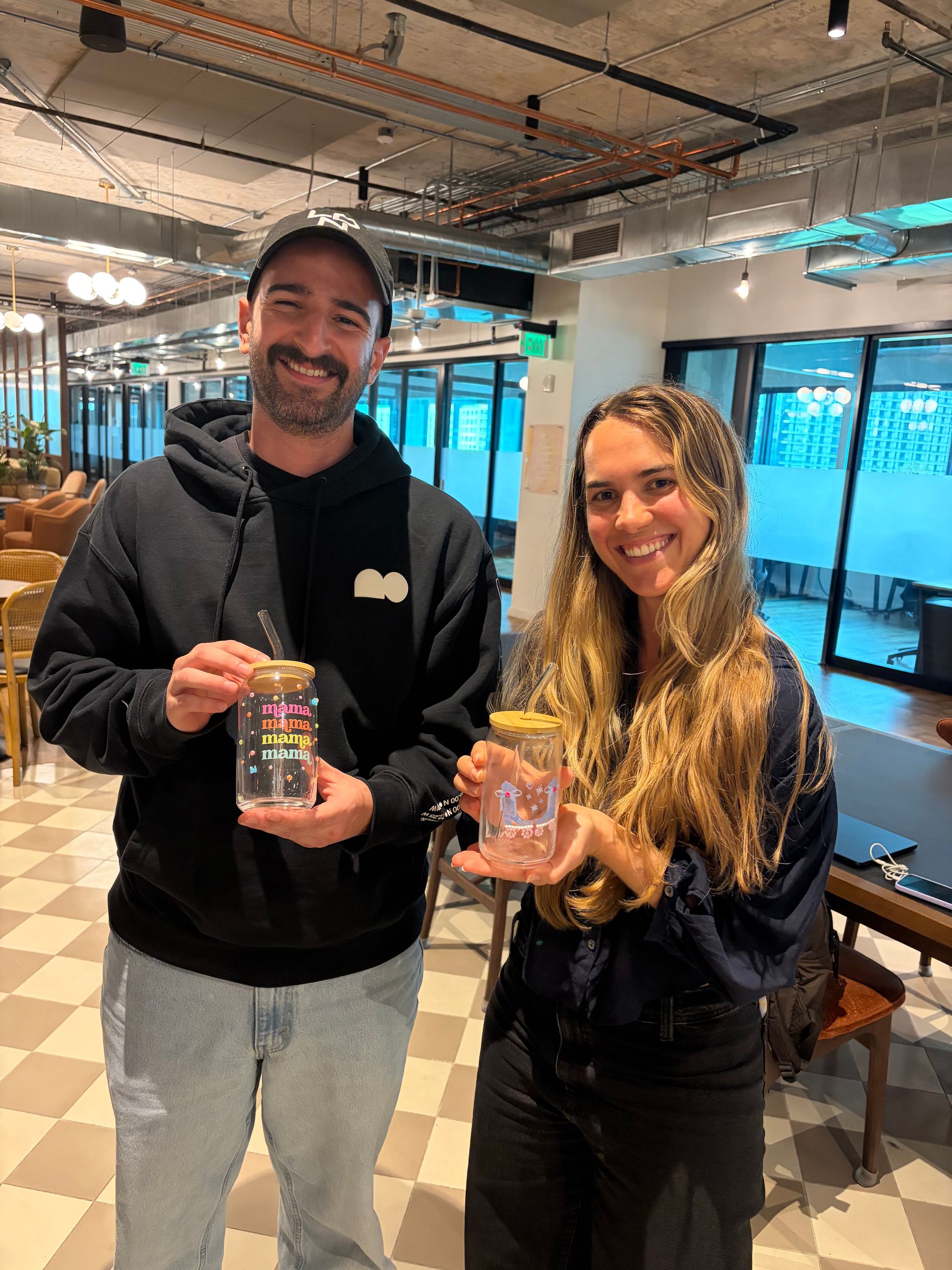 Two smiling colleagues in a modern open-plan coworking office holding decorative reusable glass jars with wooden lids and straws, exposed ductwork and lounge seating in the background.