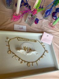 Seashell-inspired gold charm necklace with pearl accents displayed in a gift tray next to a pink velvet pouch and textured glass vases holding neon faux foliage and candles on a pastel event table.