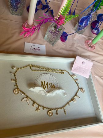 Seashell-inspired gold charm necklace with pearl accents displayed in a gift tray next to a pink velvet pouch and textured glass vases holding neon faux foliage and candles on a pastel event table.