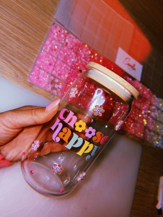 Close-up of a hand holding a clear glass jar with a bamboo lid and colorful "choose happy" lettering, decorated with tiny flower gems, over a sparkly pink glitter craft tray on a wooden table.