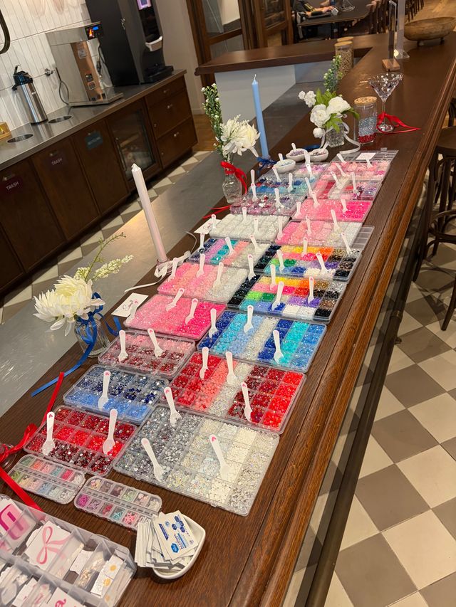 Colorful bead bar display on a wooden counter: dozens of organized plastic trays of beads and rhinestones with small scoops, decorative flowers and candles on a tiled-floor craft station.