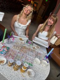 Two smiling women in white tops at an indoor artisan jewelry pop-up holding trays of necklaces above a table filled with gold chains, beads, shells and colorful candles — handmade jewelry display in a cozy boutique setting.