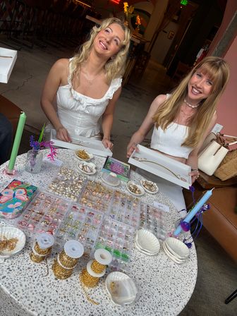 Two smiling women in white tops at an indoor artisan jewelry pop-up holding trays of necklaces above a table filled with gold chains, beads, shells and colorful candles — handmade jewelry display in a cozy boutique setting.