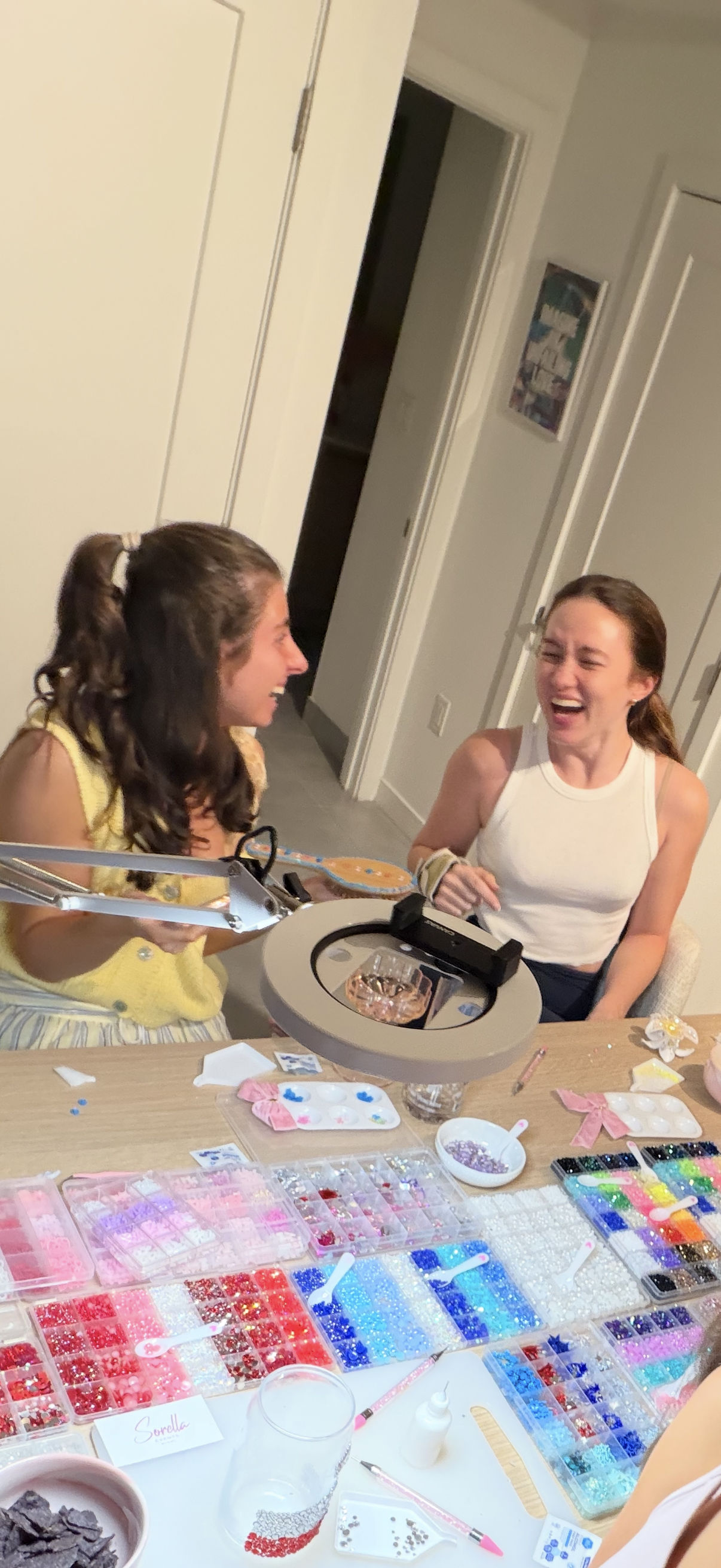 Two friends laughing at a home craft table filled with organized trays of colorful beads, rhinestones, glitter and tools, a ring light above as they work on DIY jewelry and decorations.