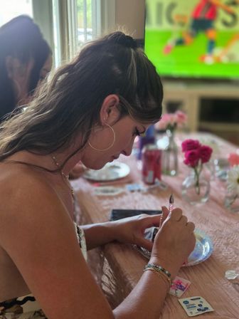 Person concentrating on a small DIY craft at a pink-decorated indoor table, using a sparkly tool to decorate a plate with vases of roses and drinks on the table and a TV in the background.