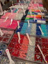 Sparkling craft beads and rhinestones organized in clear plastic trays with white scoops — rows of pink, red, blue and iridescent gems for jewelry and nail art