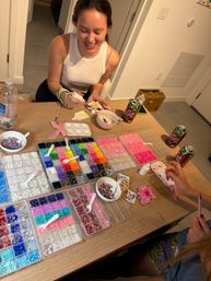 Smiling person at a home craft table gluing a flower-shaped decoration, surrounded by organized trays of colorful rhinestones, beads, sequins, bowls and craft tools for a DIY sparkle project.