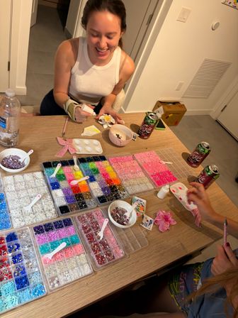 Smiling person at a home craft table gluing a flower-shaped decoration, surrounded by organized trays of colorful rhinestones, beads, sequins, bowls and craft tools for a DIY sparkle project.