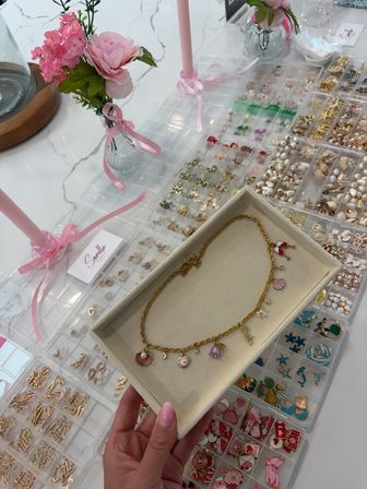 Close-up of a gold charm necklace in a cream tray held above organized clear trays of pastel enamel charms, pearls and gold findings on a white marble table with pink flowers and ribbon accents.