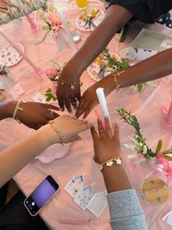 Four hands wearing rings and gold charm bracelets over a pink, floral-decorated table with taper candles, nail art, phone and small trays of beads — a festive bracelet-making party setup.