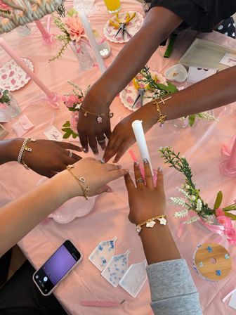 Four hands wearing rings and gold charm bracelets over a pink, floral-decorated table with taper candles, nail art, phone and small trays of beads — a festive bracelet-making party setup.